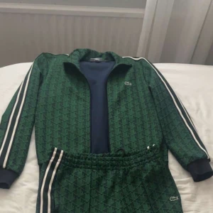 Grön dress från Lacoste med mönster - Säljer en gröna dress från Lacoste med diskret geometriskt mönster och vita ränder längs sidorna. Byxorna har resår i midjan och mudd vid bensluten samt Lacoste-logga på benet samma med koftan.