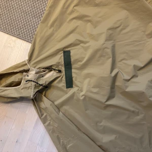 Beige regnponcho från Mero Mero - Säljer en beige regnponcho från Mero Mero med huva och justerbar dragsko. Perfekt för regniga dagar och enkel att slänga på sig över andra kläder. Stort och rymligt plagg med minimalistisk design.