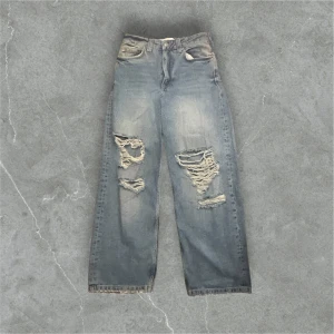 Grå slitna jeans från Bershka - Säljer ett par grå jeans från Bershka med rejält slitna detaljer på båda benen. Jeansen har rak passform och klassisk femficksdesign. Perfekta för en avslappnad och trendig look.