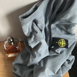 Ljusblå långärmad tröja från Stone Island - Säljer en ljusblå långärmad tröja från Stone Island med den klassiska logotypen på ärmen. Tröjan har en avslappnad passform och är tillverkad i mjuk bomull. Perfekt för en stilren och bekväm look.