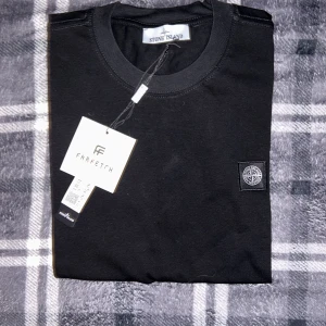 Svart t-shirt från Stone Island - Säljer en svart t-shirt från Stone Island med klassisk rund hals och ikonisk logotyp-patch på bröstet. Tröjan är i mjuk bomull och har en stilren, enkel design. Perfekt för dig som gillar streetwear och vill ha något snyggt och bekvämt.