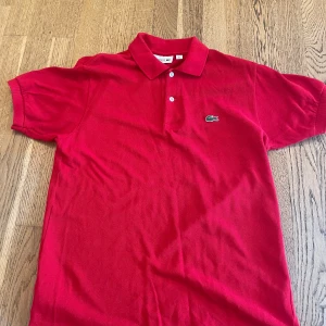 Röd pikétröja från Lacoste - Säljer en klassisk röd pikétröja från Lacoste med den ikoniska krokodilloggan på bröstet. Tröjan har krage, två knappar och korta ärmar. Perfekt för en stilren och sportig look.