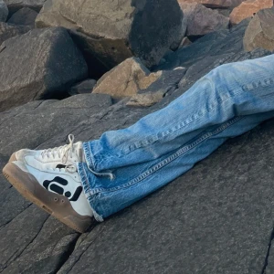 Vita sneakers med svart mönster och brun sula - Säljer ett par vita sneakers med svart grafiskt mönster på sidan och brun sula. Skorna har snörning och en klassisk rund tå. Perfekta för dig som gillar en unik stil och vill sticka ut lite extra.