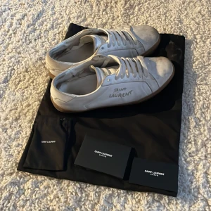 Vita sneakers från Saint Laurent - Säljer ett par klassiska vita sneakers från Saint Laurent med bruna gummisulor och snörning. Skorna har en enkel och stilren design med Saint Laurent-logga på sidan. Perfekta för dig som gillar exklusiva och tidlösa sneakers.