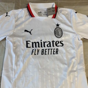 Vit AC Milan fotbollströja från Puma - Säljer en vit AC Milan fotbollströja från Puma med svart logga och klubbmärke på bröstet. Tröjan har korta ärmar, röd och svart detalj på kragen samt trycket 'Emirates Fly Better' på framsidan. Materialet är lätt och andas, perfekt för match eller träning.