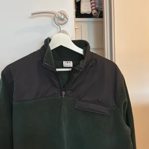 Mörkgrön fleece-tröja från Zara - Mörkgrön fleece-tröja från Zara med svarta detaljer över axlar och bröst. Tröjan har halv dragkedja framtill och en liten ficka på bröstet. Perfekt för kyligare dagar och enkel att matcha med jeans eller joggers.