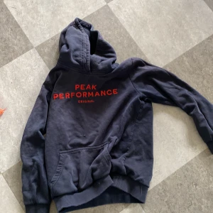 Mörkblå hoodie från Peak Performance - Mörkblå hoodie från Peak Performance med röd logga på bröstet. Klassisk huva och magficka framtill. Perfekt för en avslappnad stil.