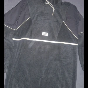 Svart fleece klänning från Reebok - Säljer en svart hoodie från Reebok i mjuk fleece med vita detaljer och dragsko i huvan. Tröjan har långa ärmar och en sportig look med Reebok-logga framtill. Perfekt för kyliga dagar eller som ett extra lager.