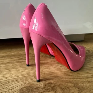 Rosa pumps med hög klack - Säljer ett par glansiga rosa pumps med spetsig tå och hög klack. Skorna har en klassisk och elegant form med lackad yta och röd sula. Perfekta för dig som vill sticka ut med färg. Oanvända storlek 36