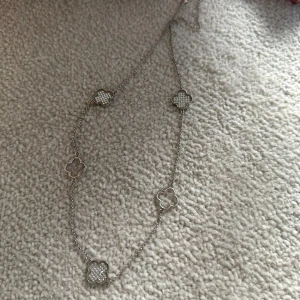 Silverfärgat halsband med fyrklöverhängen - Säljer ett elegant halsband i silverfärg med flera fyrklöverformade hängen, vissa med glittrande stenar och andra i enkel metall. Perfekt för att ge din outfit en lyxig touch.