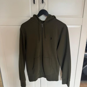 Olivgrön hoodie från Polo Ralph Lauren - Säljer en olivgrön hoodie från Polo Ralph Lauren med dragkedja och huva. Tröjan har en broderad logga på bröstet och praktiska fickor framtill. Perfekt för en avslappnad stil. Kvitto finns, Använd 4-5 gånger. Byxorna är M. PM vid frågor. 