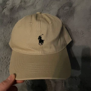 Beige keps från Polo Ralph Lauren - Snygg beige keps från Polo Ralph Lauren med klassisk broderad logga framtill och justerbart spänne bak. Perfekt accessoar för en stilren look. Märket syns även invändigt och baktill på kepsen.