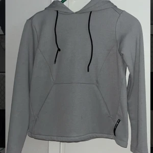 Grå hoodie från Sail Racing - Säljer en stilren grå hoodie från Sail Racing med svarta dragsnören och logga på sidan. Tröjan har en stor huva och långa ärmar, perfekt för en avslappnad look. Passar dig som gillar sportig och enkel stil.