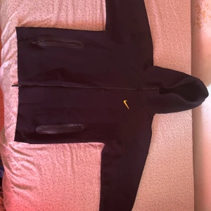 Svart kofta från Nike NOCTA - Säljer en svart hoodie från Nike NOCTA med dragkedja och huva. Tröjan har gula detaljer, två fickor med dragkedja och broderad Nike-logga på bröstet. Perfekt för dig som gillar streetwear och sportig stil. Passar bra för M också, pris kan diskuteras.