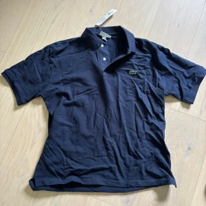 Mörkblå pikétröja från Lacoste - Säljer en klassisk mörkblå pikétröja från Lacoste med den ikoniska krokodilloggan på bröstet. Tröjan har krage, knappar vid halsen och är kortärmad. Perfekt för en stilren och sportig look.