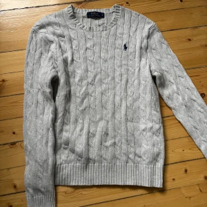 Grå stickad tröja från Polo Ralph Lauren strl S - Kabelstickad grå tröja från Polo Ralph Lauren med rund halsringning och broderad marinblå logga på bröstet. Klassisk och tidlös design med ribbade muddar vid ärmslut och nederkant. 