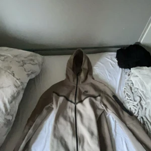 Beige fleecejacka med huva från Nike - Säljer en beige fleecejacka från Nike med svart logga på bröstet och dragkedja hela vägen. Jackan har huva och svarta detaljer längs bröstet och ärmarna. Perfekt för kyliga dagar och enkel att matcha. Passar s och xs 