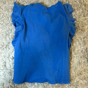 Blå ribbad topp med volangärmar - Säljer en blå ribbad topp med volangdetaljer längs ärmhålen. Toppen är ärmlös och har en mjuk, stretchig känsla. Perfekt för att addera lite färg till din outfit!