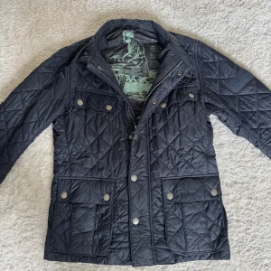 Barbour jacka - Säljer en svart quiltad jacka från Barbour med klassiskt rutmönster. Jackan har dragkedja och tryckknappar framtill samt flera fickor med lock. Perfekt för kyligare dagar och har en snygg detalj med Barbour-tryck på insidan. Storlek S