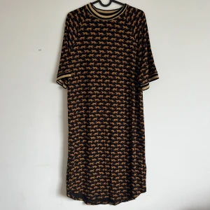 Svart klänning med tigertryck från Monki - Svart klänning från Monki med heltryck av orange tigrar. Klänningen har rund hals med guldfärgade ränder och halvlånga ärmar med matchande kant. Rak passform och mjukt material, perfekt för en cool och avslappnad look.