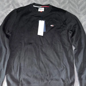 Svart stickad tröja från Tommy Jeans - Säljer en svart stickad tröja från Tommy Jeans med rund halsringning och diskret logga på bröstet. Tröjan har långa ärmar och ribbade muddar vid ärmslut och nederkant. Perfekt för en stilren och enkel look.