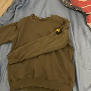Olivgrön sweatshirt från Stone Island - Säljer en olivgrön sweatshirt från Stone Island med klassisk logga på ärmen. Tröjan har rund halsringning och ribbade muddar vid ärmslut och nederkant. Perfekt för en avslappnad stil.