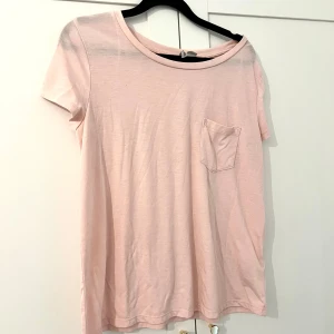 Ljusrosa t-shirt med bröstficka - En basic ljusrosa t-shirt med rund halsringning och en liten bröstficka på vänster sida. T-shirten har korta ärmar och är tillverkad i ett mjukt material som passar perfekt till vardags.