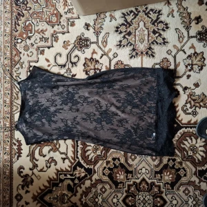 Svart spetsklänning med blommönster - Säljer en svart ärmlös klänning med transparent spets och blommigt mönster. Klänningen har en rak passform och är dekorerad med spetsdetaljer längs nederkanten. Perfekt för dig som gillar en elegant och stilren look.