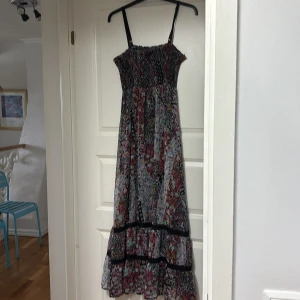 Blommig långklänning från Twentyone - Säljer en långklänning från Twentyone med smock upptill och tunna axelband. Klänningen har ett färgglatt blommigt mönster i svart, rött, blått och vitt samt volangdetaljer nertill. Perfekt för varma dagar och sommarens alla tillfällen.