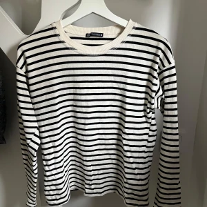 Randig långärmad tröja från Zara - Säljer en vit långärmad tröja med svarta horisontella ränder från Zara. Tröjan har rund halsringning med ribbad kant och är tillverkad i ett mjukt bomullsmaterial. Perfekt för en avslappnad och stilren look.