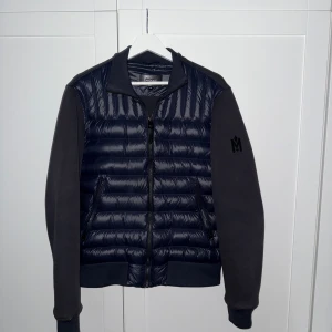 Mackage cardigan - Säljer en snygg blå hybridjacka från Mackage med quiltad, glansig front och ärmar i mjukare tyg. Jackan har dragkedja och en diskret logga på ärmen. Perfekt för dig som vill ha en stilren och sportig look.