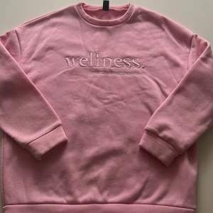 Rosa sweatshirt med broderad text från SHEIN - Mysig rosa sweatshirt från SHEIN med broderad text 'wellness. INVEST IN YOUR HAPPINESS' på bröstet. Tröjan har rund halsringning och långa ärmar, perfekt för en avslappnad stil. Använd ett fåtal gånger men har blivit lite knopprig. Lite av trådarna har gått upp🩷