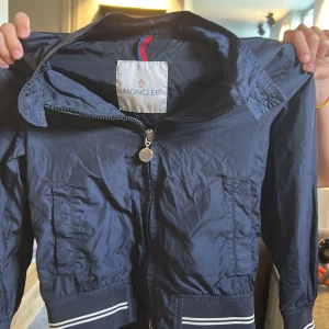 Mörkblå vindjacka från Moncler - Säljer en mörkblå vindjacka från Moncler med dragkedja framtill och ribbade muddar med vita ränder. Jackan har två fickor och klassisk Moncler-logga på ärmen. Perfekt för blåsiga dagar och snygg till vardags.fråga om ni funderar över något