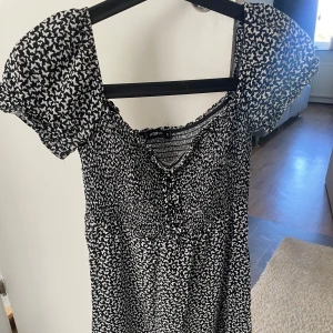 Svartvit blommig klänning från FB Sister - Supersöt svartvit klänning från FB Sister med puffärmar och smock upptill. Klänningen har ett blommigt mönster och en liten v-ringning med knyt framtill. Perfekt för dig som gillar en feminin och trendig stil.