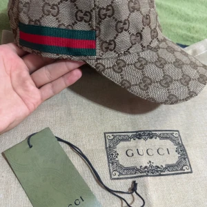 Gucci beige keps med GG-mönster - Snygg beige keps från Gucci med klassiskt GG-monogram över hela kepsen och en grön-röd rand på sidan. Tillverkad i Italien med exklusiv känsla och detaljer i brunt skinn. Perfekt för dig som vill ha en lyxig och trendig accessoar.priser kan diskuteras!😊