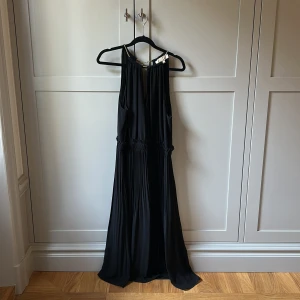 Svart långklänning från Michael Kors - Supersnygg svart långklänning från Michael Kors med plisserad kjol och guldfärgade detaljer vid halsen. Klänningen är ärmlös och har en elegant, flowy siluett med nyckelhålsöppning fram. Slutar över knän för mig som är 165cm!