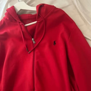 Röd hoodie från Polo Ralph Lauren - Röd hoodie från Polo Ralph Lauren med svart broderad logga på bröstet. Tröjan har huva med snörning, dragkedja framtill och praktiska fickor. Perfekt för en chill och stilren look.