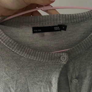 Grå kofta från 157 med knappar - En basic grå kofta från 157 med rund halsringning och knäppning framtill. Långärmad modell i mjuk bomullsblandning, perfekt att slänga över en t-shirt eller linne. Ribbstickade muddar vid ärmslut och nederkant för en clean look.