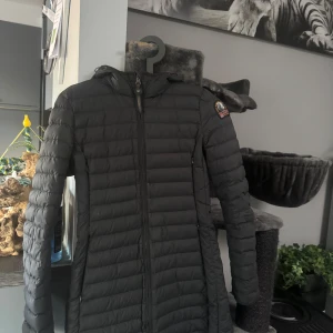 Svart lång dunjacka med huva - Säljer en svart lång dunjacka från Parajumpers med huva och dragkedja framtill. Jackan har quiltad design, två fickor med dragkedja och en patch på ärmen. Perfekt för kalla dagar när du vill hålla dig varm och snygg.