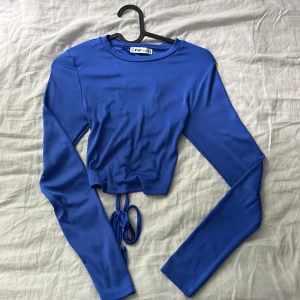 Blå croppad topp med snörning FB Sister - Säljer en blå croppad långärmad topp från FB Sister. Toppen har snygg snörning och öppen rygg baktill som ger en cool och trendig look. Tillverkad i ett stretchigt syntetmaterial som sitter bra. 