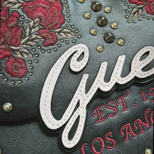 Svart väska från Guess - Snygg svart väska från Guess med broderade röda rosor, nitar och stora logotypdetaljer. Väskan har både handtag och axelrem, samt dragkedja inuti. ❤️🌹skriv om ni har frågor!!