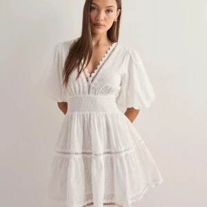 Vit kortklänning med volang och spets - Superfin vit kortklänning med v-ringning och puffärmar,  Klänningen har volangdetaljer, spets vid halsringningen och ett diskret prickigt mönster. Perfekt för sommarens alla tillfällen! Nypris 2500kr