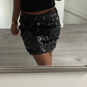 Svart paljettkjol med glans - Säljer en svart kortkjol täckt med stora glittriga paljetter som ger en riktigt festlig vibe. Kjolen har en tight passform och är perfekt för dig som vill sticka ut med lite extra bling. Supersnygg att matcha med en enkel topp och boots.