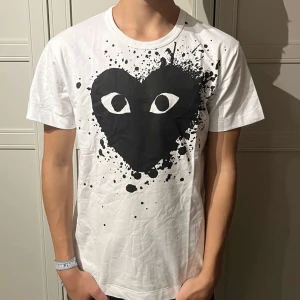 Vit t-shirt Comme des Garçons Play - Säljer en vit t-shirt från Comme des Garçons Play med stort svart hjärta och ögon på bröstet, omgiven av coola färgstänk. Klassisk rund hals och korta ärmar. Perfekt statement-plagg för dig som gillar streetstyle och unika prints. (Helt ny)!🤝
