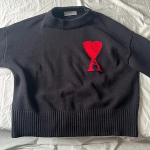 Svart stickad tröja från Ami med hjärta - Svart stickad tröja från Ami med ribbade muddar och hög krage. På bröstet finns ett stort rött hjärta med bokstaven A, vilket ger en unik och trendig look. Perfekt för dig som gillar stilrena plagg med en twist.