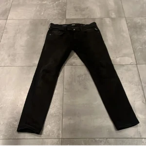 Svarta Replay Hyperflex jeans - Säljer ett par svarta Replay Hyperflex Anbass jeans med slim passform. De har klassisk femficksdesign, svarta sömmar och stretchigt material som sitter skönt. Perfekta om du gillar stilrena och bekväma jeans med snygg look.jeansen är lite solbrända vilket har gett dom en brun look men det är inget märkvärdigt