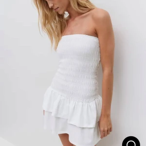 Vit axelbandslös kort klänning - Supersöt vit klänning med smock upptill och volang i nederdelen. Axelbandslös design. Klänningen är köpt från Gina Tricot för 400kr. Använt en gång. Skriv för fler frågor och funderingar.