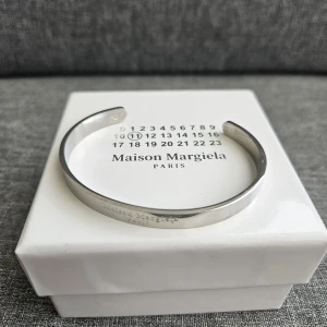 Maison Margiela silverarmband - Stilrent och minimalistiskt armband från Maison Margiela. Armbandet har en öppen design och är graverat med märkets logga. Perfekt accessoar för dig som gillar clean och modern stil.