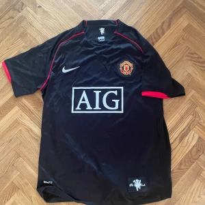 Manchester United svart Nike tröja M - Säljer en svart Manchester United fotbollströja från Nike med röd detalj längs axlar och ärmslut. Tröjan har klubbmärket på bröstet, AIG som sponsor och är tillverkad i lätt polyester med Nike FITDRY-teknologi. MUFC-tryck på ryggen.