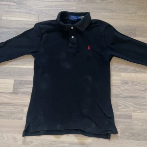 Svart pikétröja från Ralph Lauren - Svart långärmad pikétröja från Polo Ralph Lauren i slim fit. Tröjan har klassisk krage, två knappar och rött broderat Polo-logga på bröstet.  en skön  Perfekt för dig som gillar stilrena och tidlösa plagg.